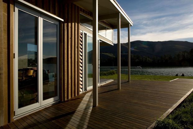 Exterior view of a contemporary lakeside building with vertical timber cladding, expansive glazed doors and windows, and a broad timber deck extending toward the water. A slender covered veranda with simple structural posts frames views of the surrounding hills and lake.