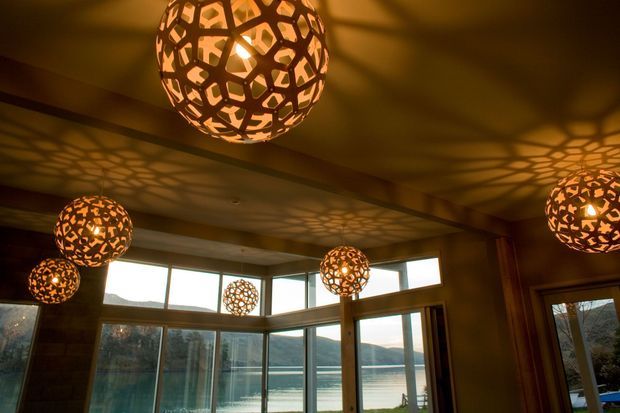A warm, moody interior with multiple spherical pendant lights featuring intricate perforated metal shades. The lights cast dramatic geometric shadows across the ceiling and walls. Large floor-to-ceiling windows and glazed doors provide views of a calm waterfront landscape outside, while exposed timber beams and dark warm-toned finishes define the room.