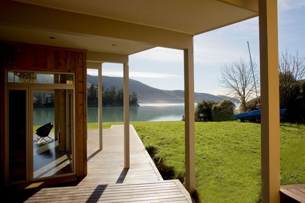 View from a covered timber deck or terrace toward a calm lake and surrounding hills. The image shows a simple open-sided roof structure supported by square posts, timber decking, a glazed timber-framed doorway, and a grassy waterfront setting. The interior beyond the doorway is only minimally visible, while the deck and lakeside outlook dominate.