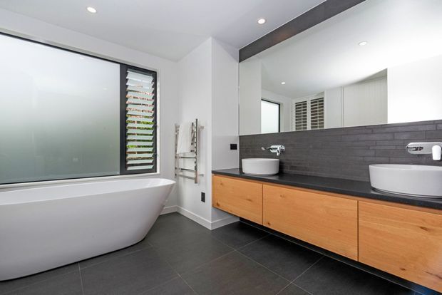 Contemporary bathroom with a freestanding white bathtub, twin vessel basins, timber vanity cabinetry, dark tiled floor and backsplash, a large wall mirror, and louvred window glazing. Recessed ceiling lights and a dark exposed ceiling beam add crisp architectural detail.