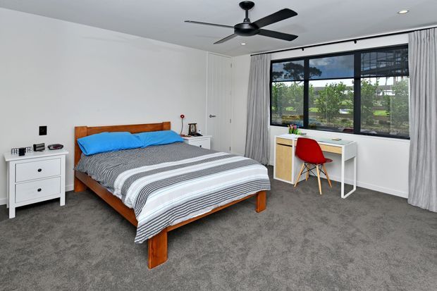 A bright, modern bedroom with a timber bed frame, grey carpet, striped grey-and-white bedding, white bedside storage, and a compact white desk beneath a large black-framed window. A red chair provides a small accent of colour, while full-height grey curtains and a black ceiling fan complete the room.