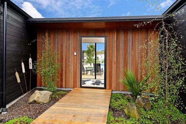 A modern courtyard entry framed by dark horizontal-clad walls, with a warm vertical timber-clad facade and central glazed door. A timber boardwalk leads through landscaped gravel, rocks, mossy groundcover and ornamental planting toward the entrance. The reflection/glazing reveals an outdoor seating area beyond, while the flat roofline and strong timber-and-black palette create a contemporary, sheltered approach.