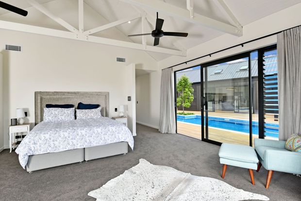 Bright, spacious bedroom with a large upholstered bed, patterned white bedding, grey carpet, bedside tables, and exposed white roof trusses with a ceiling fan. Full-height black-framed sliding glass doors open to a poolside terrace, with light curtains and two pale blue lounge chairs in the foreground.