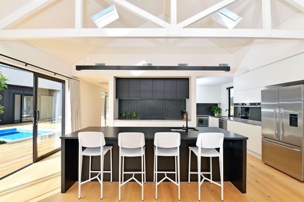 Bright, spacious contemporary kitchen with a large black island and four white bar stools, light timber flooring, white cabinetry, stainless-steel appliances, and a dark textured splashback. Exposed white roof trusses and skylights create a high, airy ceiling, while expansive glazing opens toward an outdoor pool area.