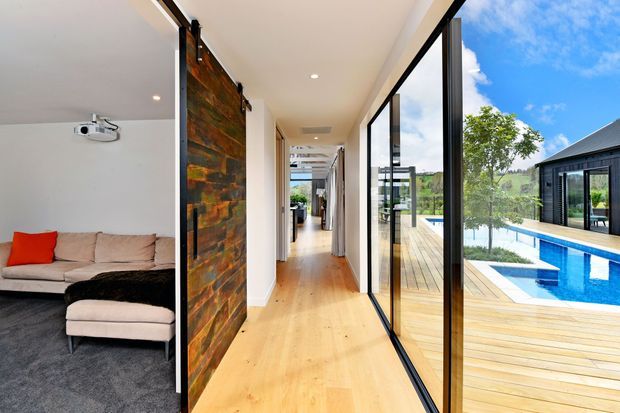 Bright contemporary interior corridor with warm timber flooring, white walls, and expansive black-framed glazing overlooking a timber deck and swimming pool. A living room with sectional sofa and projector is visible to the left, while a large sliding barn-style door clad in rustic timber forms a feature element beside the hallway.
