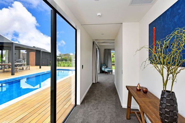 Bright contemporary interior hallway with grey carpet, white walls, and full-height black-framed glazing opening onto a timber pool deck and swimming pool. A timber console with decorative objects and a large blue artwork lines the wall, while a seating area is visible at the end of the corridor. The adjoining exterior includes a covered outdoor entertaining area and pool.