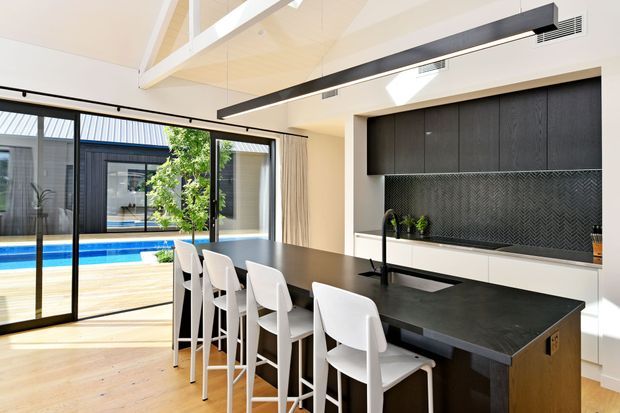 Contemporary open-plan kitchen with a large dark island and seating for five, integrated black cabinetry, a textured herringbone splashback, and expansive sliding glass doors opening toward a timber deck and swimming pool. The room has pale timber flooring, white walls and ceiling beams, strong natural light, and a long linear pendant light.