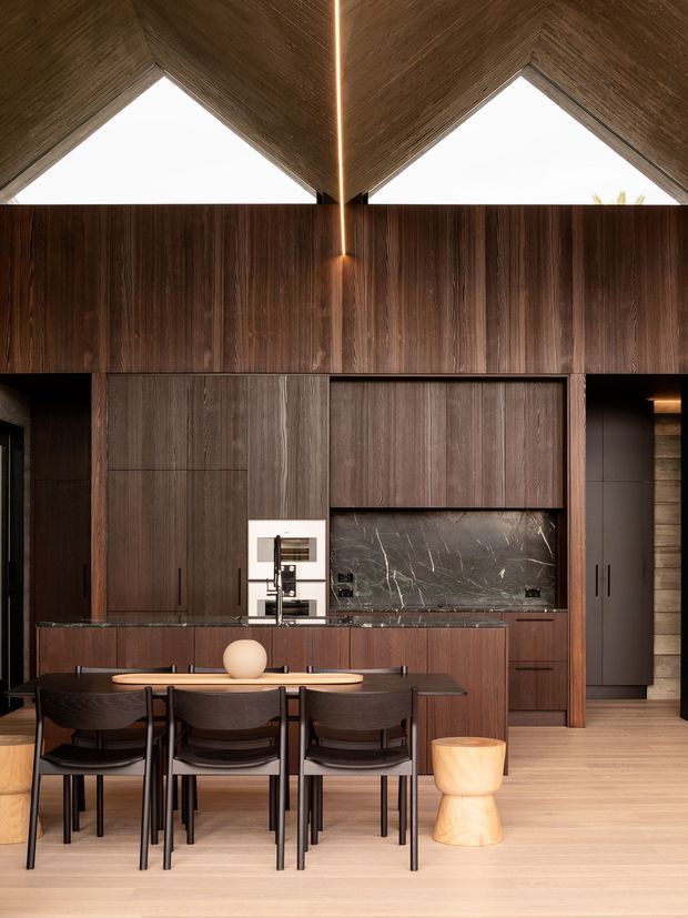 A sophisticated open-plan kitchen and dining area with extensive dark timber cabinetry, a dark veined stone splashback and benchtops, and a central island facing a dining table for four. The room has a dramatic pitched timber-lined ceiling with triangular clerestory glazing and a slim linear pendant or concealed light at the ridge. Warm timber flooring, sculptural dining chairs, and a small pale timber stool create a refined, moody interior.