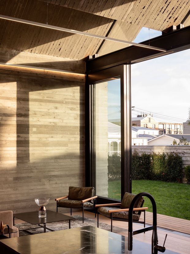 A refined modern interior with exposed board-formed concrete walls and a dramatically angled concrete ceiling. Full-height dark-framed glazing opens views to a small lawn, timber deck and neighbouring urban buildings. Leather-and-wood lounge chairs, a low dark coffee table, linear pendant lighting and a reflective stone island or benchtop create a warm, sculptural living space with strong indoor–outdoor connection.