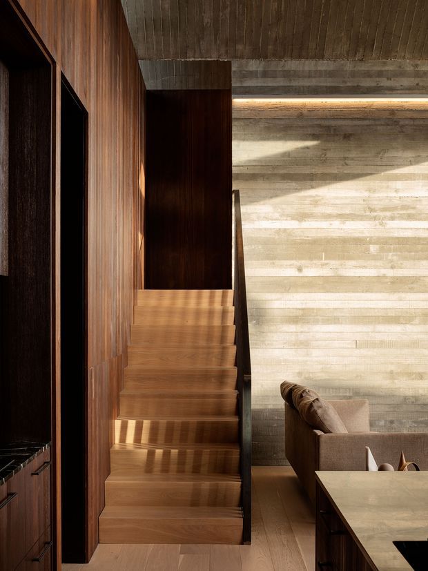 A dramatic interior view centered on a sculptural timber staircase rising beside a dark timber-panelled wall. The staircase and flooring use warm natural wood, while a horizontally textured pale concrete or timber feature wall is illuminated by concealed linear lighting. Part of a contemporary living or dining area with a neutral upholstered sofa and stone-like tabletop is visible at right. Strong contrast between dark joinery, warm timber, pale textured wall surfaces, and directional sunlight creates a refined, moody atmosphere.