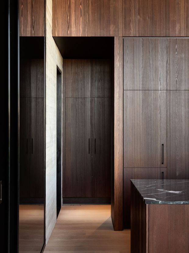 A close-up interior view of a dark, refined built-in cabinetry installation. Floor-to-ceiling timber-grain wardrobes and storage panels frame a narrow passage, with slim black handles and a dark stone benchtop visible in the foreground. A pale stone-clad vertical partition provides contrast against the rich wood finishes and warm timber flooring. The image emphasises bespoke joinery, material texture, and a moody contemporary aesthetic.