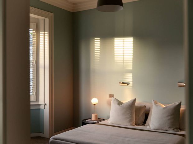 A softly lit bedroom with a neatly made bed, layered neutral cushions, muted blue-green walls, white decorative cornice, and warm timber-toned trim. Plantation shutters cast striped sunlight onto the wall, while a small bedside lamp and wall-mounted reading lights add a calm, refined atmosphere.