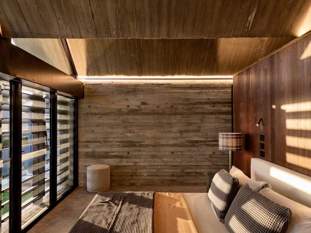 A warm, compact contemporary bedroom with extensive timber finishes. The room features a timber-lined ceiling, horizontally clad timber feature wall, dark timber wall paneling, integrated bedside controls, a low upholstered bed with patterned cushions, a small upholstered ottoman, and full-height louvred glazing allowing striped sunlight into the space. Recessed linear lighting and earthy brown tones create an intimate, refined atmosphere.