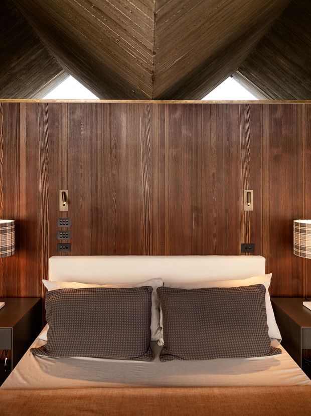 A close-up, symmetrical view of a sophisticated bedroom focused on the bed wall. The bed has a cream upholstered headboard, dark textured cushions, and warm neutral bedding. Rich dark timber wall panelling rises behind the bed, with integrated bedside electrical outlets and slim wall lights. Matching dark bedside tables and cylindrical fabric lamps flank the bed. Above, a dramatic timber-lined pitched or folded ceiling creates a strong architectural focal point.