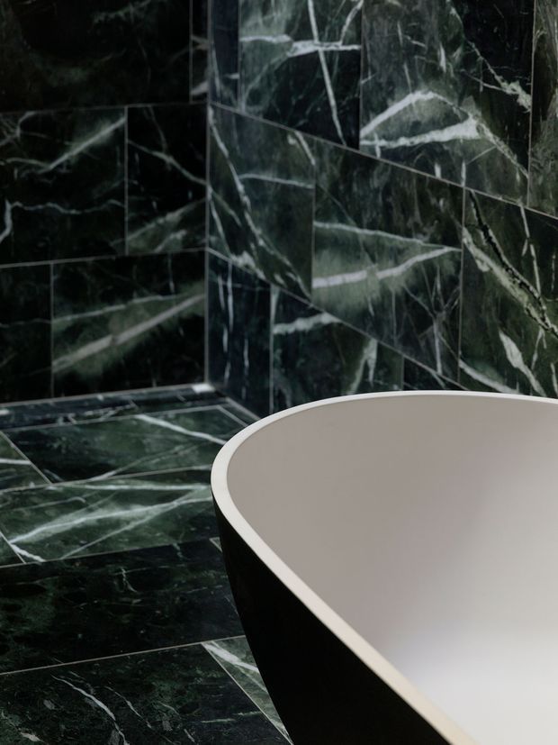 Close-up view of a luxurious bathroom featuring a freestanding bathtub with a pale interior and dark exterior, set against dramatic dark green-black marble-look tiled walls and floor with bold white veining. The image is tightly framed around the bath and marble surfaces, creating a moody, high-end atmosphere.