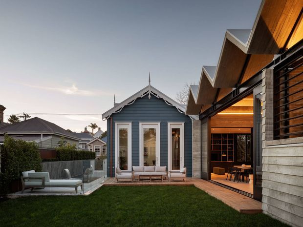 A landscaped residential courtyard viewed from outdoors, featuring a restored-looking blue weatherboard cottage with ornate gable trim and white-framed windows, connected visually to a contemporary extension on the right. The extension has a strongly articulated zigzag roofline, warm timber soffits, horizontal timber cladding, black-framed glazed openings, and an open dining/living area. A lawn, timber decking, outdoor seating, hedges, and neighbouring suburban homes complete the scene at dusk.