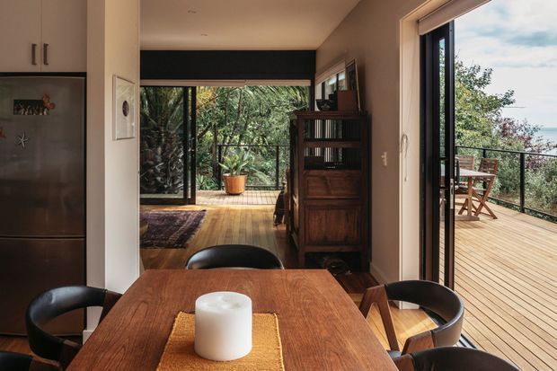 A warm, compact open-plan interior with a timber dining table and black curved chairs in the foreground. A dark built-in kitchen with stainless-steel refrigerator occupies the left side, while the rear living area features timber flooring, a vintage wooden bookcase, and extensive black-framed glazing. Folding or sliding glass doors open onto a timber deck with outdoor dining, surrounded by dense greenery and glimpses of water beyond.