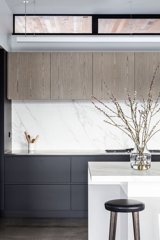 Contemporary kitchen with dark lower cabinetry, textured grey wood-grain upper cabinets, a white marble-look backsplash, and a white island with a matching veined surface. Linear pendant lights and decorative wood panels add architectural interest, while a black leather bar stool and vase of branches provide minimal styling.