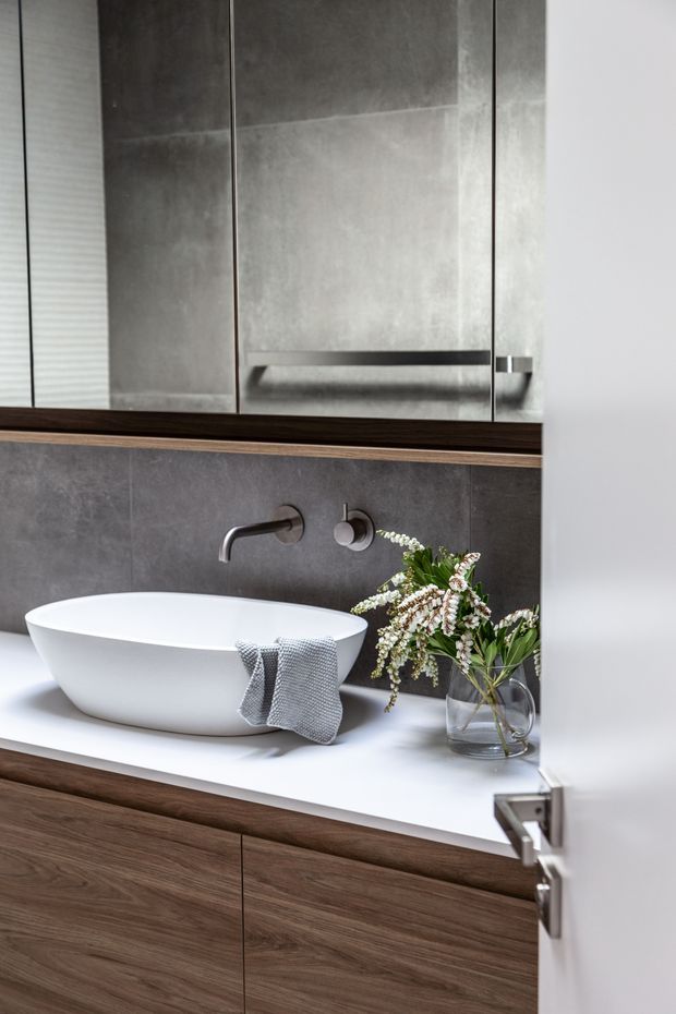 Contemporary bathroom vanity featuring a white vessel basin on a white benchtop, wall-mounted brushed metal tapware, dark grey tiled wall surfaces, and warm timber cabinetry. Mirrored storage cabinets with integrated horizontal handles sit above the vanity, while a small glass vase and folded grey towel add styling.
