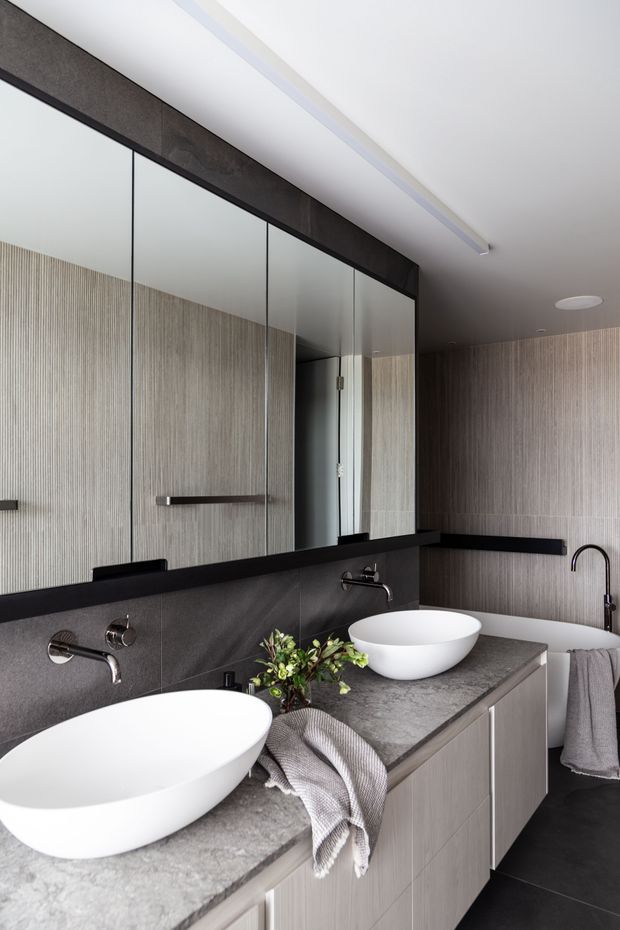 Contemporary double-vanity bathroom with two white vessel basins set on a grey stone benchtop, wall-mounted chrome tapware, pale vertically textured wall tiles, dark grey wall cladding and flooring, a large mirrored wall, and light timber-toned built-in vanity cabinetry. A freestanding white bathtub is partially visible at the right.