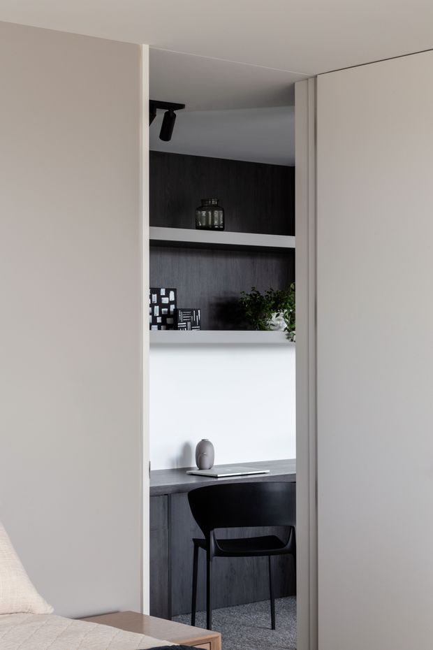 View from a bedroom through an open doorway into a compact built-in home office. The office features a dark timber-look desk and shelving, white walls and joinery, a black chair, understated decorative objects, and a small plant. The bedroom foreground includes a light upholstered bed and timber bedside surface. The overall design is restrained and monochromatic with strong black, white, and dark wood contrasts.