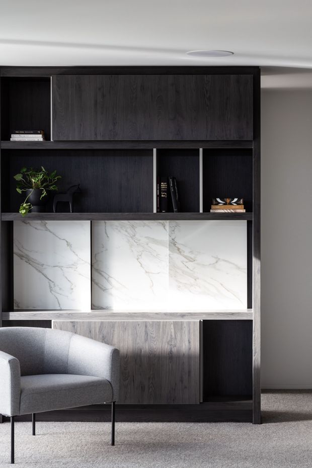 A refined built-in dark timber shelving and cabinetry unit fills the frame, with open display niches, books, a potted plant, and a large white marble-look feature panel. A grey upholstered armchair sits in front on a neutral carpet, creating a quiet, contemporary reading or living area.