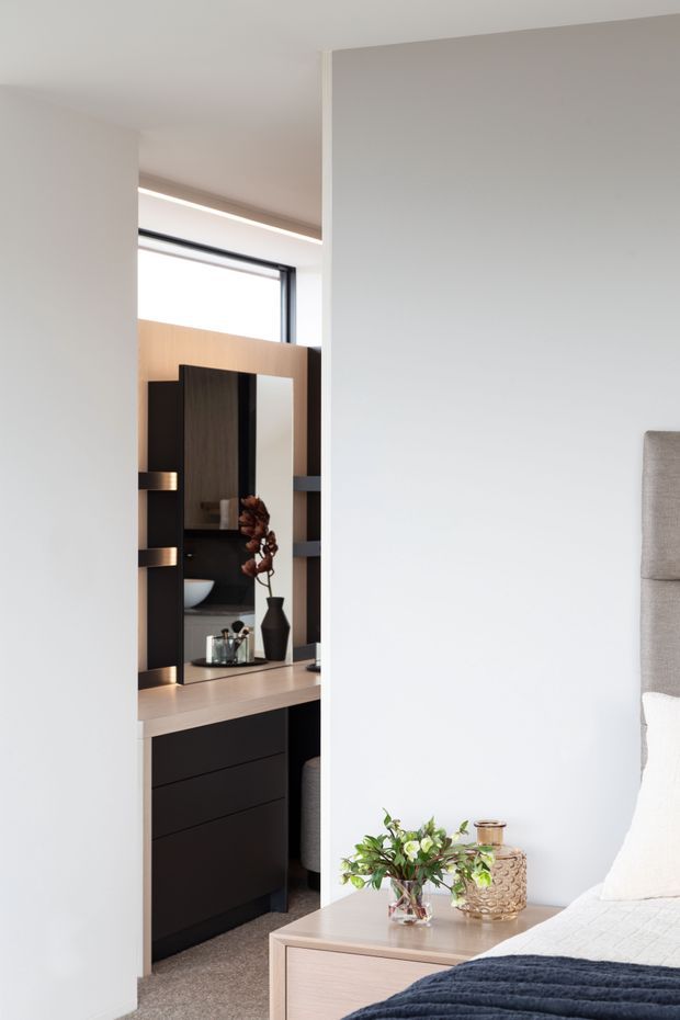 A bright, contemporary bedroom viewed toward a compact built-in dressing and vanity area. The room has white walls, a pale timber bedside table, upholstered bedhead, dark navy bedding, and a dark timber and black built-in joinery unit with a mirror, drawers, shelves, and integrated lighting. Neutral finishes and carefully styled accessories create a refined, calm interior.