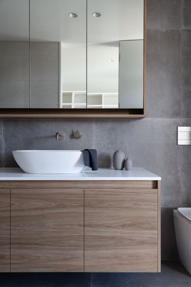 A refined contemporary bathroom featuring a floating timber-look vanity with a white countertop and freestanding vessel basin. Large grey wall tiles create a subdued backdrop, complemented by a wide mirrored cabinet with a timber frame, brushed metal wall-mounted tapware, and minimal neutral accessories. A partial toilet is visible at the right edge.
