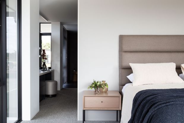 A contemporary bedroom with a large upholstered taupe-grey bedhead, white bedding, and a dark navy textured throw. A compact timber bedside table with a small plant sits beside the bed. Full-height glazing and a narrow hallway or dressing area are visible to the left, with a restrained neutral palette and dark accents.