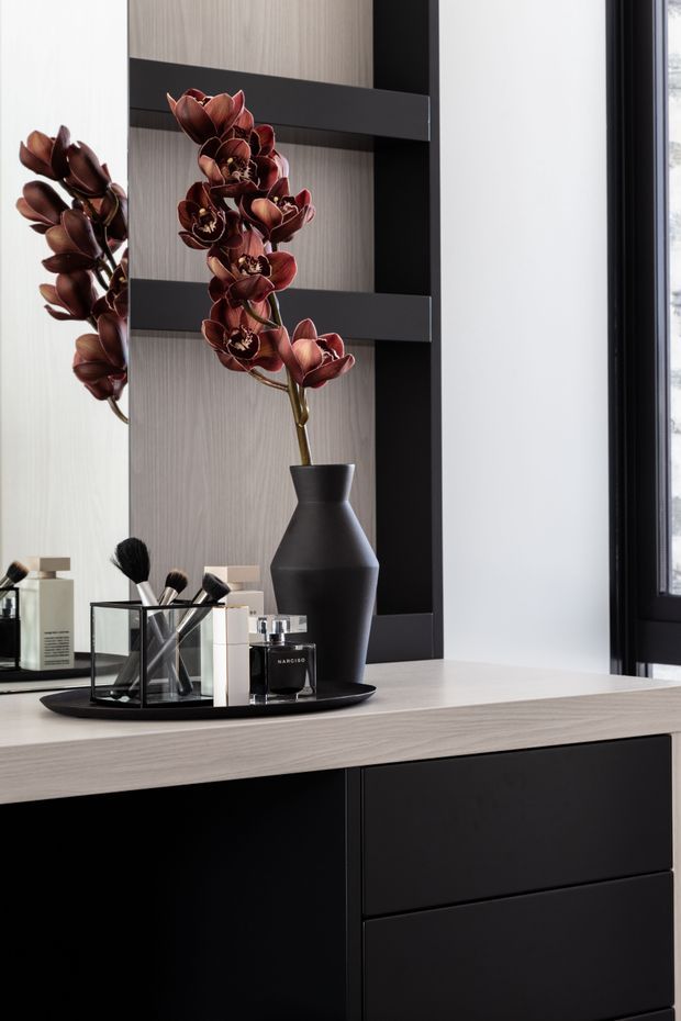 Close-up of a refined vanity or dressing area featuring a pale timber-look countertop and matching vertical wall panel, matte black drawer cabinetry, and a black-framed open shelving niche. A large mirror reflects the floral arrangement, while a dark sculptural vase, cosmetics, and makeup brushes sit on a black tray. A black-framed window is visible at right, providing natural light.