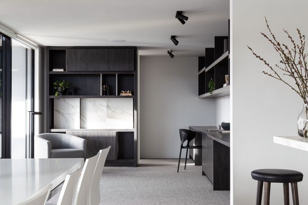 Contemporary open-plan interior combining a dining area, living space and compact kitchen. The room features dark timber built-in cabinetry, a white marble-look benchtop or feature panel, pale walls, grey carpet, black track lighting, and large glazed openings. A white dining table with chairs occupies the foreground, while a dark kitchen counter and stools appear to the right.