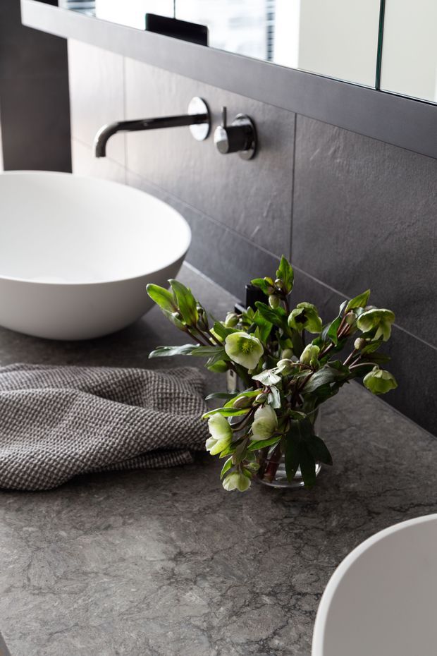 Close-up view of a contemporary bathroom vanity with a dark grey stone countertop, white vessel basins, a wall-mounted black tap, grey tiled splashback, and a large mirror. A folded textured towel and floral arrangement add softness to the otherwise monochromatic scheme.