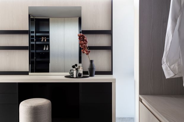 A refined dressing and wardrobe area with a dark built-in vanity desk, pale stone or composite benchtop, upholstered stool, open shelving, and a large central mirror. Integrated wardrobe cabinetry is visible on the right, with neutral wood-grain finishes, black joinery, and carefully styled accessories.