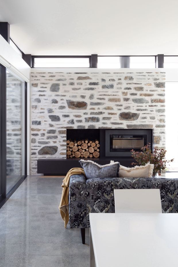 Bright contemporary living room with a large exposed rough-stone feature wall, integrated dark fireplace and log niche, expansive black-framed glazing, polished grey concrete floor, and a patterned sofa facing a white dining table and chairs. The space combines strong natural materials with a refined, modern interior.
