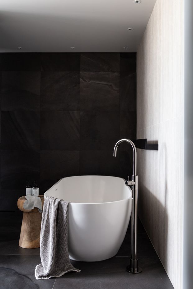 A sophisticated contemporary bathroom featuring a freestanding white oval bathtub, tall chrome floor-mounted faucet, dark stone-look wall and floor tiles, and a vertically textured light wall surface. Soft towels and a small wooden stool add warmth, while recessed ceiling lights and strong side lighting create a moody, refined atmosphere.