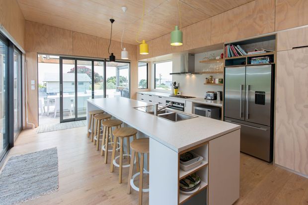 Bright open-plan kitchen with a long white island and integrated sink, seating for several people on timber stools, extensive plywood wall and ceiling lining, timber flooring, stainless-steel appliances, open shelving, and colourful pendant lights. Large glazed sliding doors connect the kitchen to an outdoor balcony or terrace.