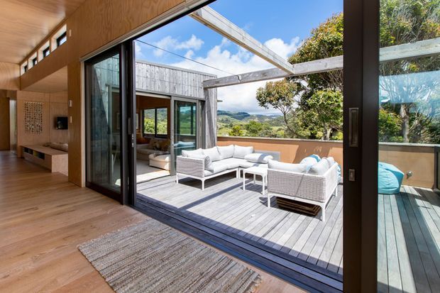 A warm timber-lined interior opens through large black-framed sliding glass doors onto a sunny timber deck. The outdoor terrace has white wicker lounge seating, a glass or open pergola structure, and views across a green, hilly landscape. A built-in timber media or display unit and woven rug are visible inside.
