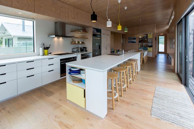 A bright, elongated contemporary kitchen with a large white island and integrated seating for several people. White lower cabinetry lines the cooking wall, while extensive warm timber cabinetry and a timber ceiling create a cohesive finish. The room includes stainless-steel appliances, open shelving, pendant lights in varied colours, pale timber flooring, and extensive glazing along one side.