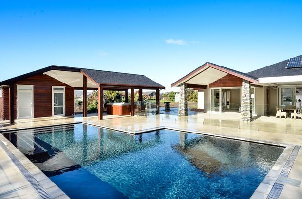 A modern single-storey pavilion-style residence arranged around a large rectangular swimming pool. The composition shows timber-clad wings with pitched tiled roofs, a covered outdoor entertaining area, stone-clad structural columns, extensive glazing, pale paved terraces and a solar panel visible on the right-hand roof. The pool and surrounding architecture dominate the bright outdoor scene.