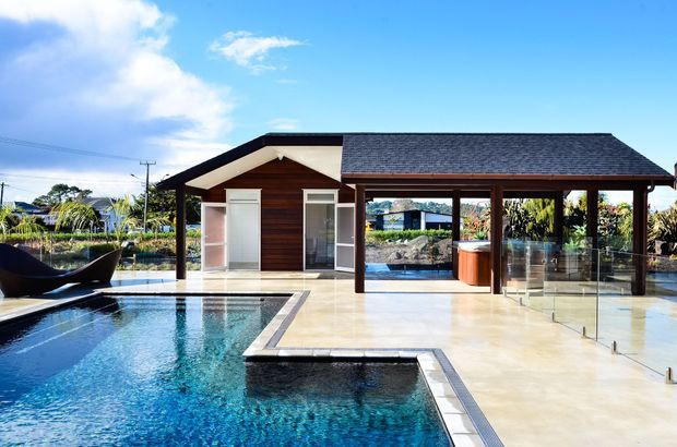 A contemporary poolside pavilion with a dark timber-clad gabled structure, glazed doors, and a covered outdoor entertaining area. The pavilion opens onto a large tiled terrace beside a swimming pool and raised spa, with transparent glass balustrading and landscaped surroundings.