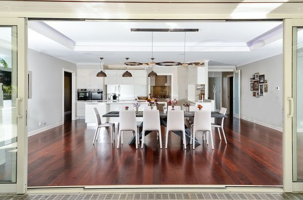 Bright open-plan dining and kitchen interior viewed through fully opened glazed folding doors. A large white dining table with upholstered chairs occupies the foreground, with a contemporary white kitchen, pendant lights, dark timber flooring, and a recessed ceiling feature beyond. The room has a clean, refined, light-filled appearance.