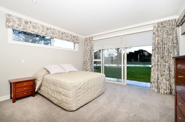 Bright, spacious bedroom with a neutral upholstered bed, beige carpet, floral patterned curtains and matching Roman blind, timber bedside table and chest of drawers. Large glazed doors and windows open the room visually to a green outdoor area.
