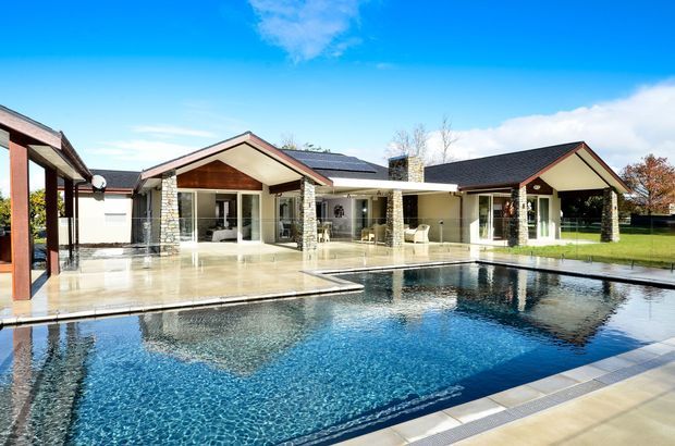 Single-storey contemporary residence arranged around a large swimming pool, with multiple low-pitched gabled wings, extensive glazed doors, stone-clad columns, light rendered walls, dark roofing and generous paved poolside terraces. The image is an exterior architectural view with a clear suburban landscaped setting.