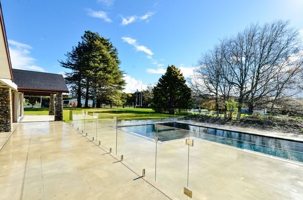 A bright outdoor pool area beside a modern house, featuring a long rectangular swimming pool, broad pale concrete paving, frameless glass safety fencing with metal fittings, and a manicured lawn with mature trees beyond. Part of the house and a stone-clad column are visible at the left edge.
