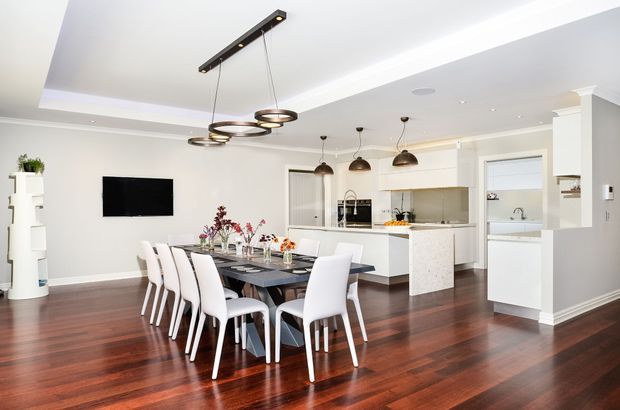 Bright open-plan contemporary interior combining a dining area and white kitchen. A long dark dining table with white upholstered chairs sits on polished dark timber flooring, beneath sculptural pendant lighting. The kitchen features white cabinetry, a light stone island benchtop, stainless-steel appliances and a pale tiled or stone splashback. Recessed ceiling details and a neutral palette create a clean, refined atmosphere.