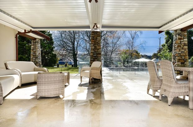 Bright, open-sided covered outdoor entertaining area with a translucent paneled roof, stone-clad columns, expansive glazing, polished pale flooring, and woven outdoor seating arranged around a lounge area. The space overlooks a landscaped suburban setting and has a relaxed indoor-outdoor feel.