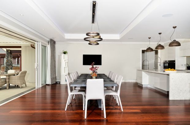 Bright open-plan dining and kitchen interior with dark polished timber flooring, a long dining table with white upholstered chairs, a white stone kitchen island, pendant lighting, and large sliding glass doors opening to an outdoor entertaining area. The room has a clean, refined contemporary fit-out with layered ceiling details and neutral finishes.