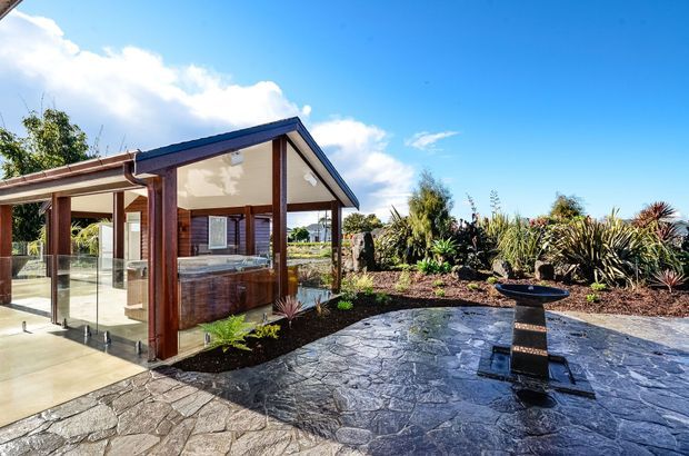 A sunny exterior view of a contemporary pavilion-style outdoor living area, with a timber-framed glazed enclosure, covered roof, outdoor kitchen or bar zone, and expansive dark irregular stone paving. A landscaped garden with sculptural grasses and succulents surrounds the entertaining space.