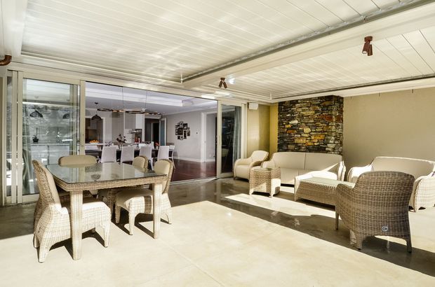 Enclosed, sunlit outdoor-style entertaining room with a dining table and woven chairs in the foreground, wicker lounge seating to the right, and a prominent stacked-stone feature wall. Large sliding glazed doors open toward an adjoining interior dining and kitchen area. The room has a white-lined ceiling, pale walls, and a polished light floor.