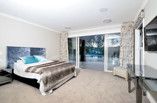 Bright, spacious contemporary bedroom with a large upholstered bed, turquoise accent pillows, a textured grey feature headboard, and a plush neutral carpet. Full-height sliding glass doors open to an outdoor pool area, with patterned curtains, a wall-mounted television, and a mirrored console contributing to the polished interior.
